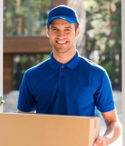 Lächelnder Umzugshelfer in blauer Uniform trägt einen Umzugskarton vor einem modernen Gebäude.